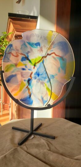 a translucent wavy circular glass plate with a smattering of various light colors swirling within. A portion of the plate has been fuse together with gold leaf.